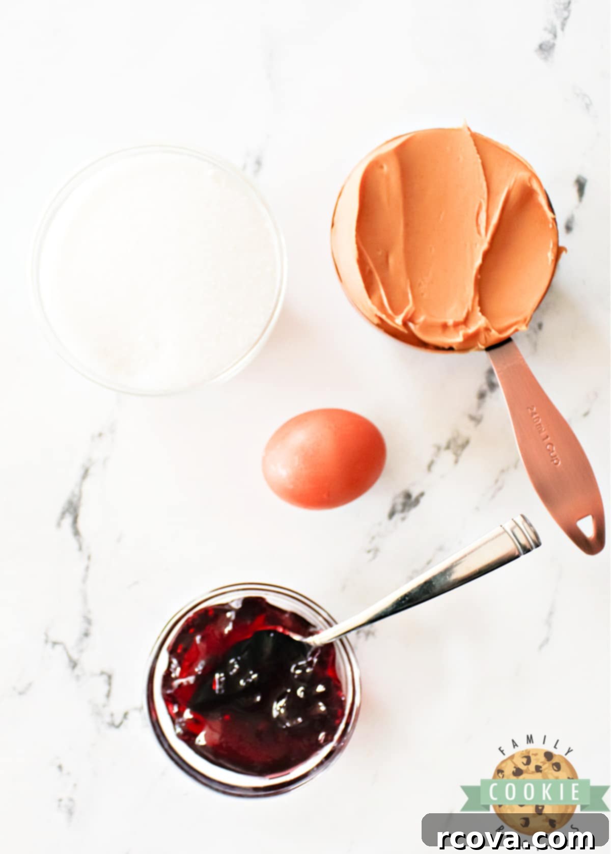 Peanut Butter Jelly Cookie Dreams 3 A flat lay image displaying the simple ingredients required for Peanut Butter and Jelly Cookies: a jar of creamy peanut butter, a bowl of granulated sugar, a single egg, and a jar of strawberry jelly.