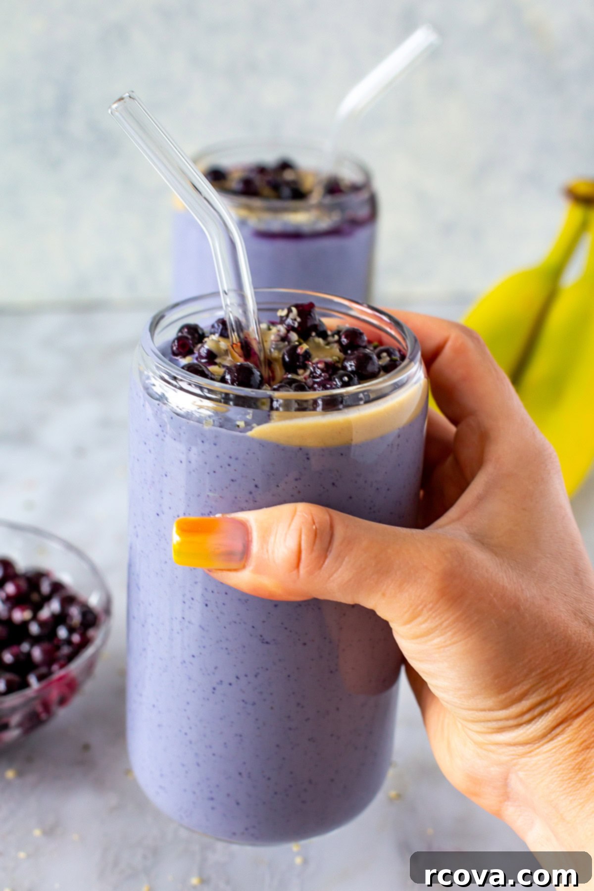 Power-Packed Protein Blends for Wellness 12 Side shot of a hand holding a purple tofu smoothie topped with blueberries and peanut butter, with two bananas in the background, a unique and healthy protein source.