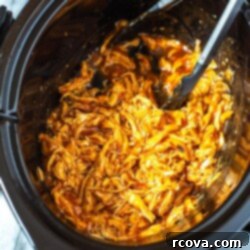 Effortless 2-Ingredient Slow Cooker BBQ Chicken 9 A close-up shot of a slow cooker filled with shredded BBQ chicken with open tongs inside.
