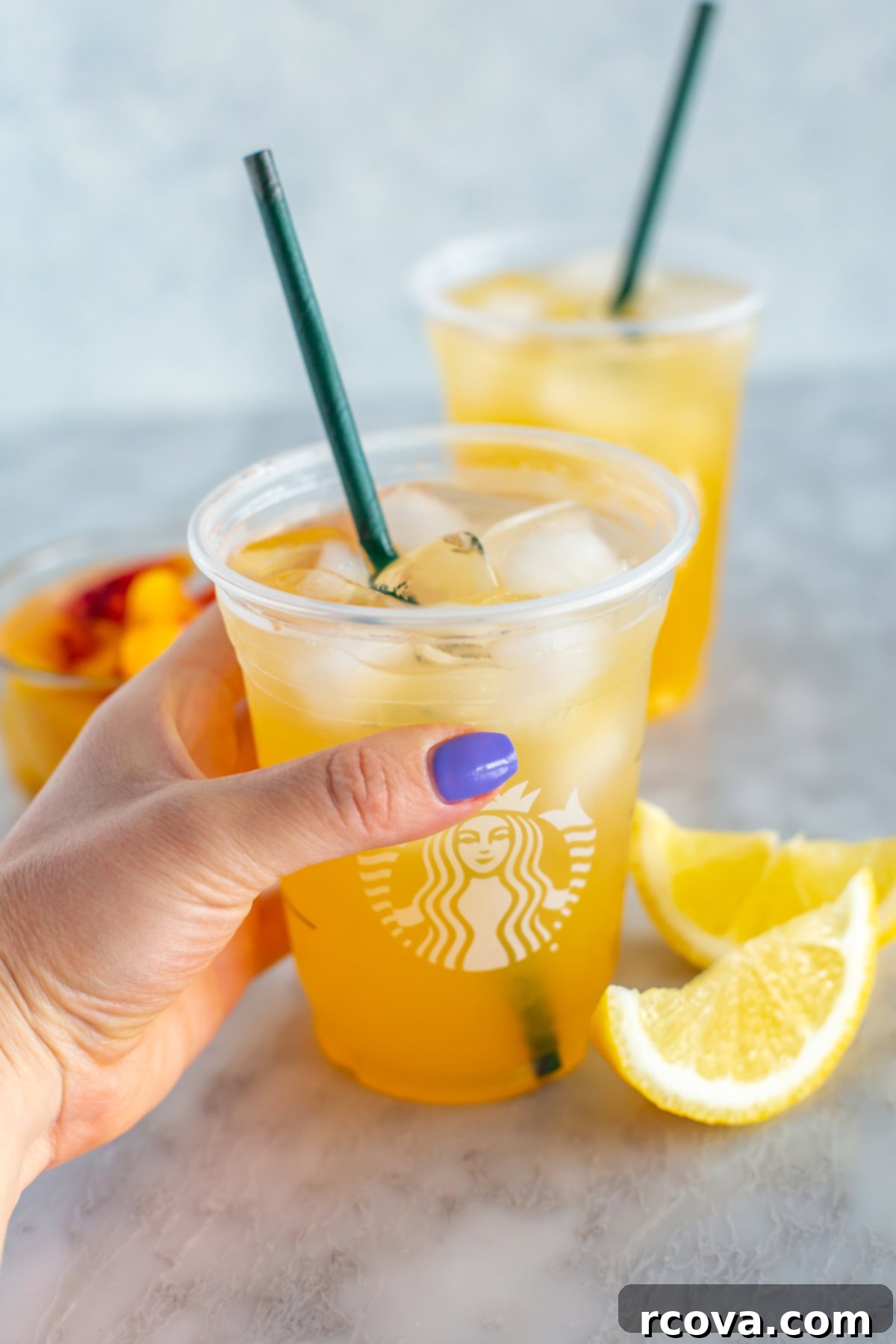 Starbucks Refreshing Iced Peach Green Tea Lemonade 8 A hand holding a clear glass of copycat Starbucks iced peach green tea lemonade with ice and a straw.