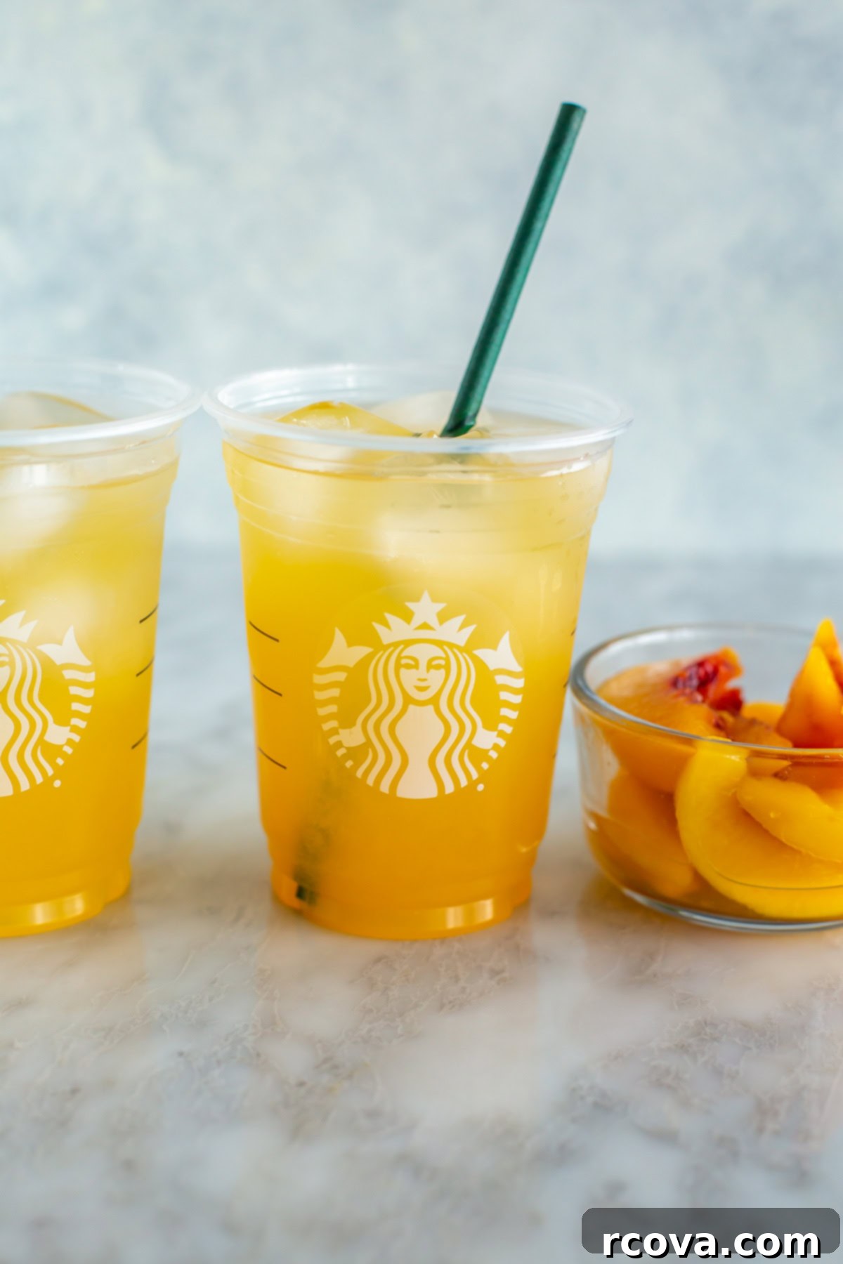 Starbucks Refreshing Iced Peach Green Tea Lemonade 9 A single clear glass of copycat Starbucks iced peach green tea lemonade with ice, peach slices, and a straw on a table.