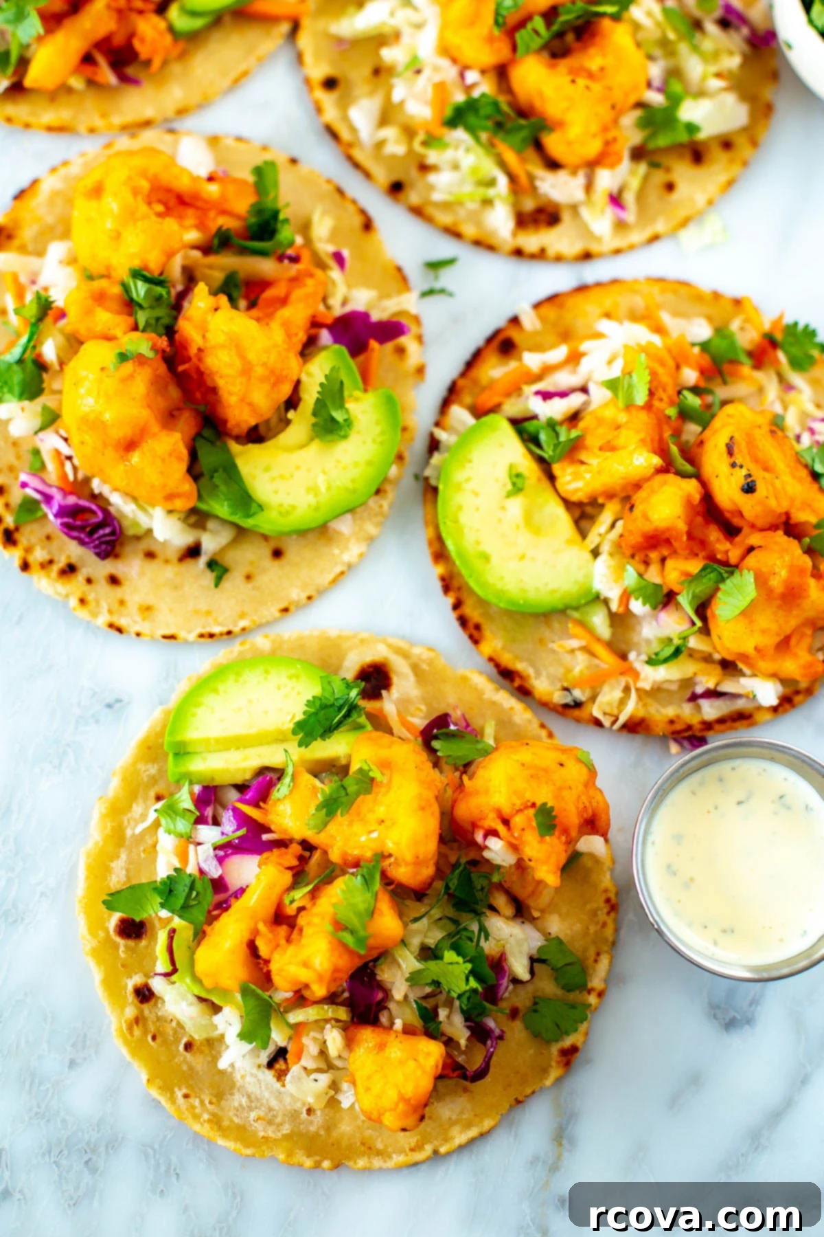 Zesty Buffalo Cauliflower Tacos 2 A close-up shot of four buffalo cauliflowe tacos topped with avocado slices with a side of ranch dressing.