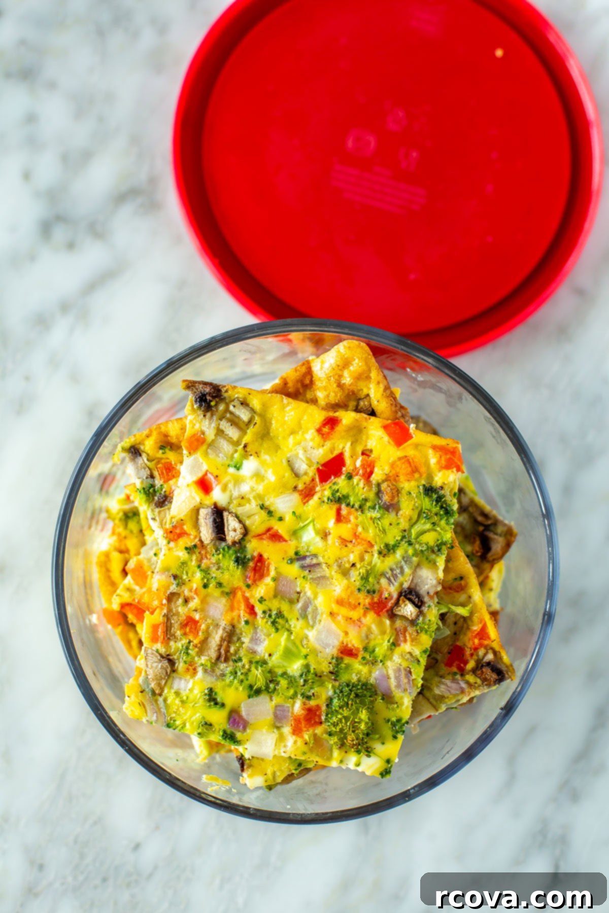 Big Batch Sheet Pan Eggs 10 Neatly cut square portions of sheet pan eggs with vegetables, stacked inside a round glass meal prep container, ready for storage.
