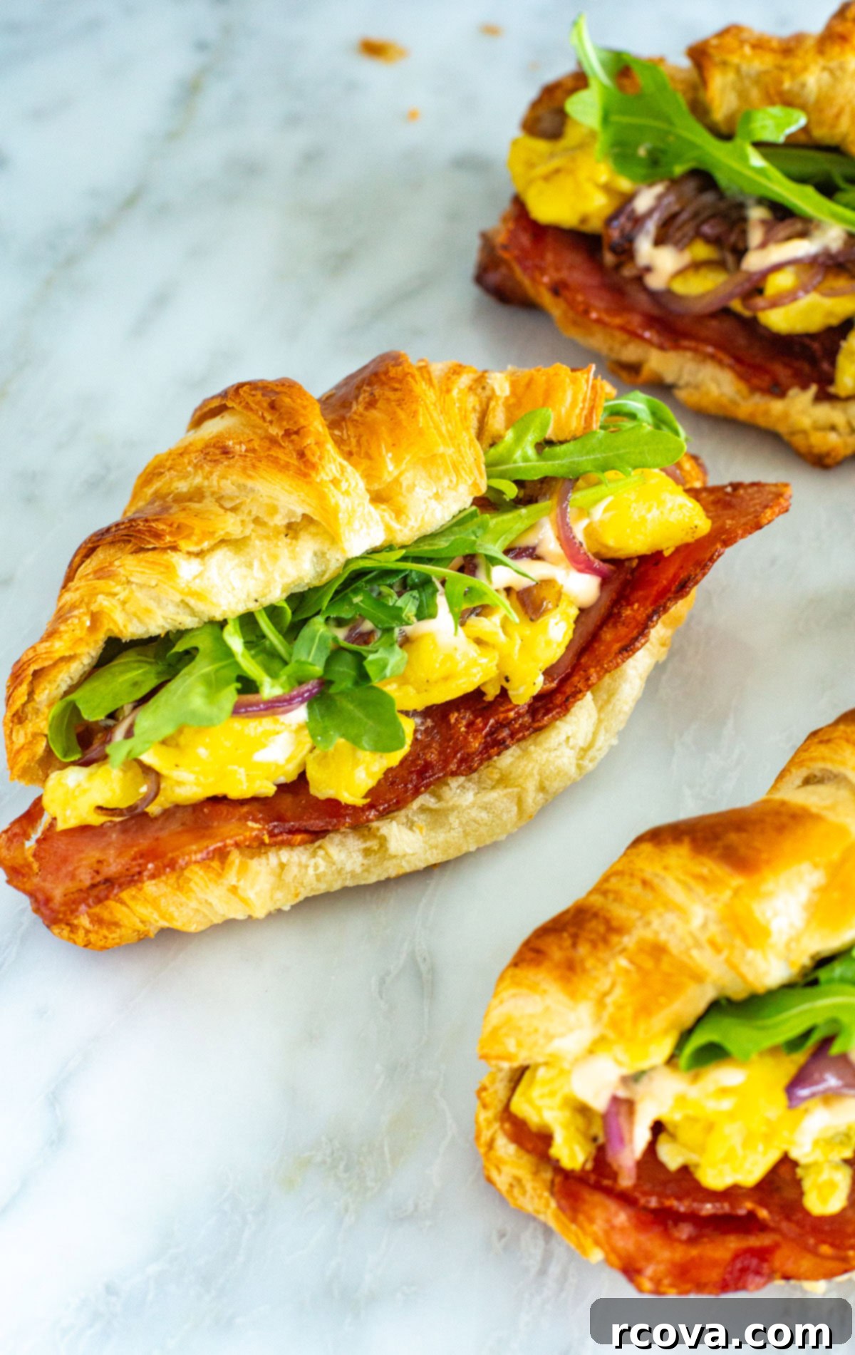 Ready-to-Go Croissant Breakfast Sandwiches 8 A tantalizing close-up shot of a single croissant breakfast sandwich, highlighting the layers of crispy bacon, fluffy scrambled eggs, fresh arugula, and hot sauce aioli spilling out slightly from the golden, flaky pastry.