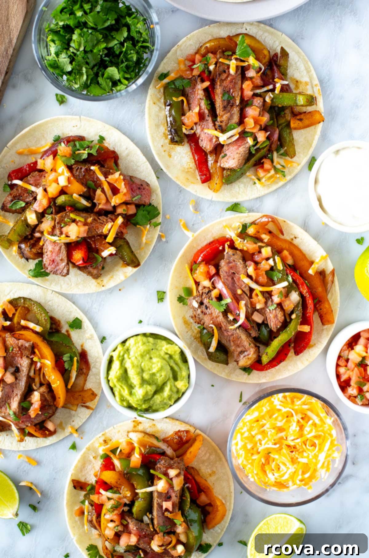 Sizzling Steak Fajitas 10 Overhead shot of a full fajita spread: assembled steak fajitas in warm tortillas are arranged with bowls of vibrant guacamole, zesty salsa, rich shredded cheese, creamy sour cream, chopped cilantro, and fresh lime wedges, ready for a customizable and delicious meal.