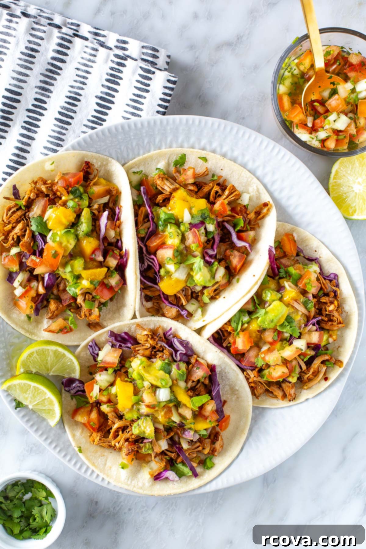 Deliciously Simple Jackfruit Tacos 2 Overhead shot of four jackfruit tacos on a white plate, garnished with shredded jackfruit, vibrant mango avocado salsa, and crisp red cabbage. Lime wedges and a small bowl of fresh cilantro are also visible on a stylish marble surface.