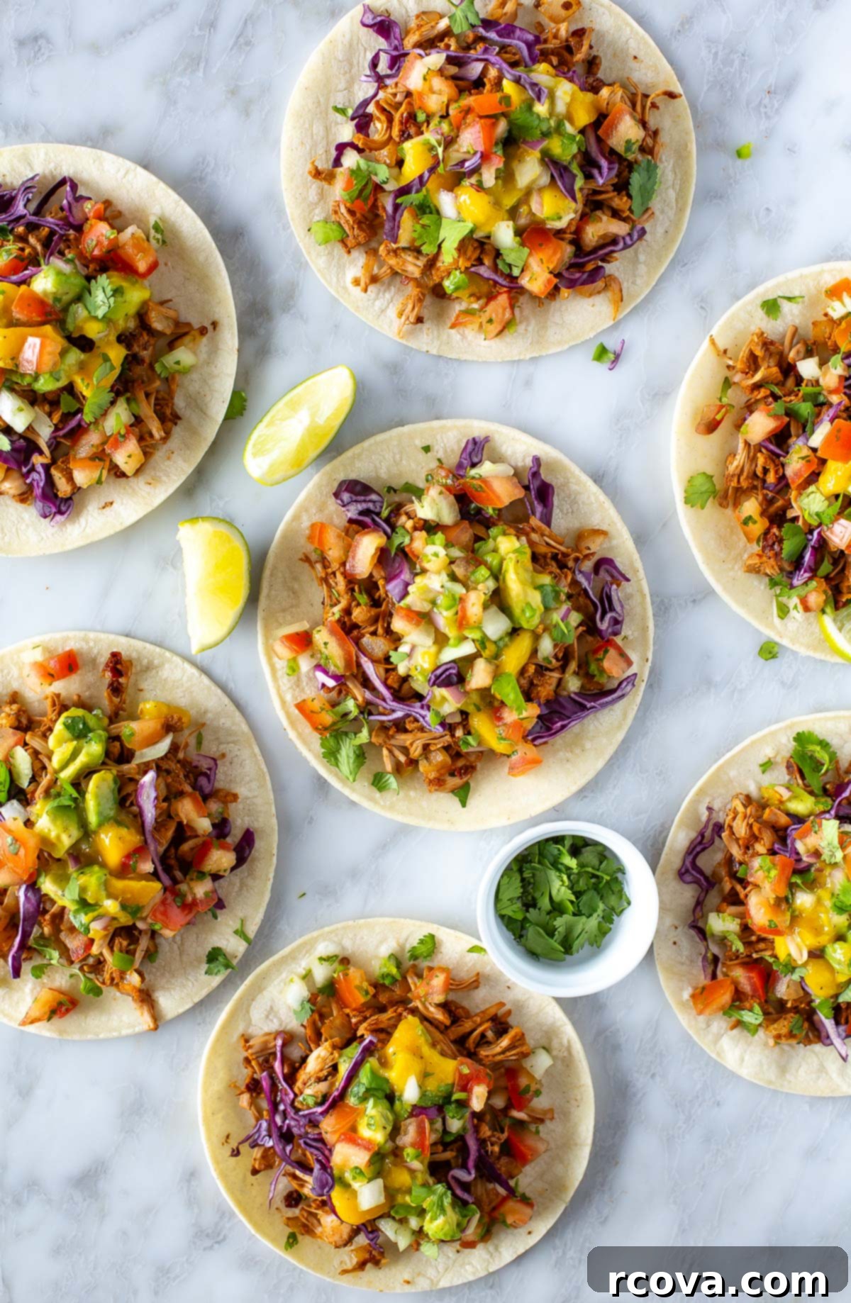 Deliciously Simple Jackfruit Tacos 8 Overhead shot of six beautifully assembled jackfruit tacos on a marble surface, each overflowing with shredded jackfruit, creamy mango avocado salsa, and thinly sliced red cabbage. Garnishings of lime wedges and a small bowl of cilantro complete the fresh presentation.