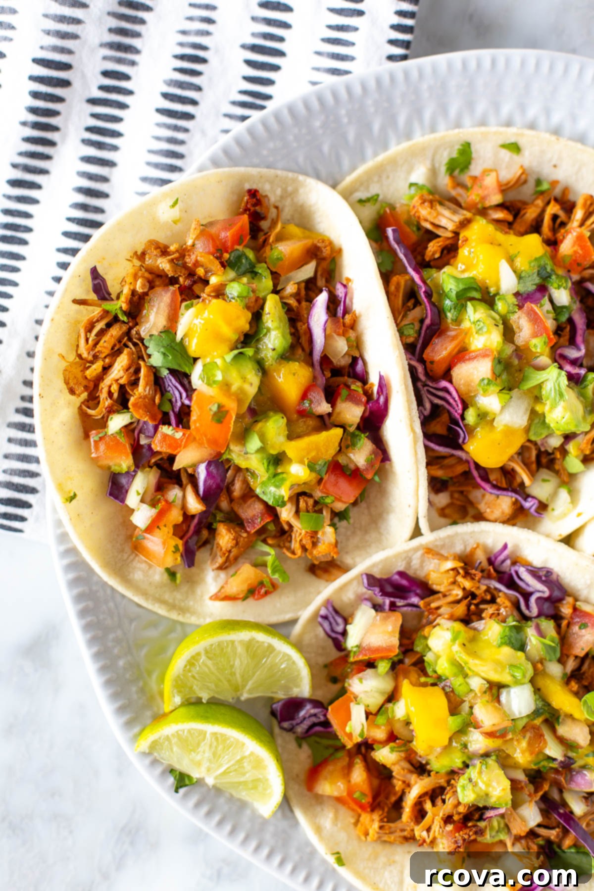 Deliciously Simple Jackfruit Tacos 9 Close-up overhead shot of three jackfruit tacos on a white plate, beautifully topped with shredded jackfruit, a generous portion of mango avocado salsa, and crisp red cabbage. Fresh lime wedges are artfully arranged on the side.