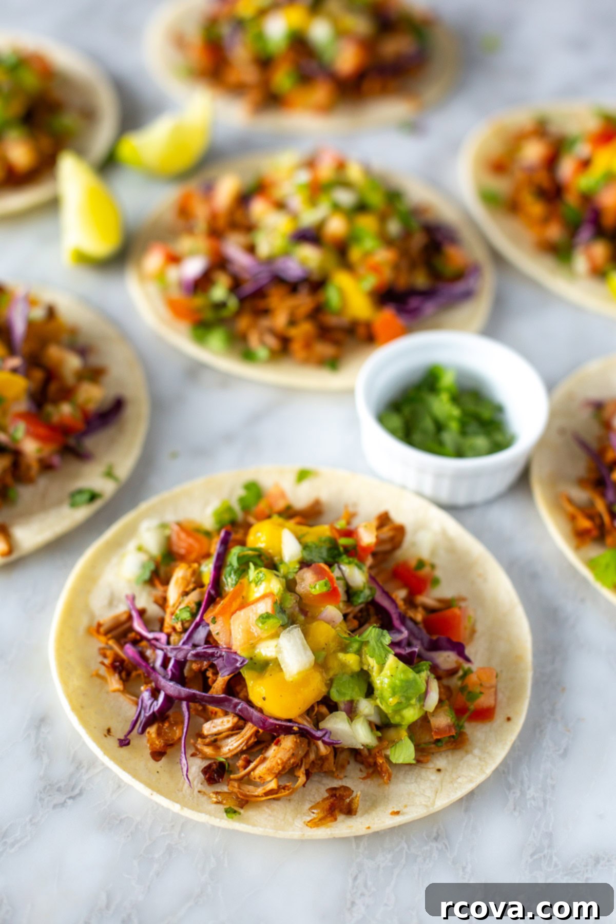 Deliciously Simple Jackfruit Tacos 10 Close-up overhead shot of several inviting jackfruit tacos displayed on a marble surface. Each taco is generously topped with shredded jackfruit, a vibrant scoop of mango avocado salsa, and fresh red cabbage. Nearby, lime wedges and a small bowl of cilantro add to the fresh appeal.