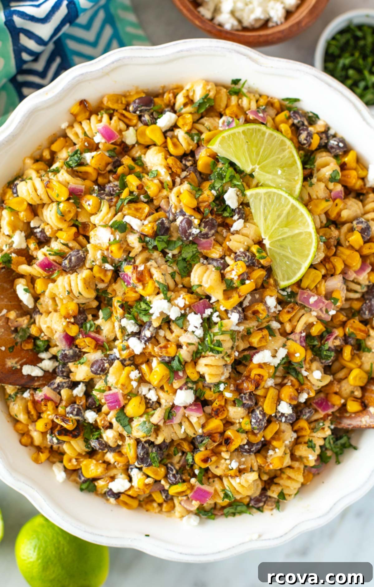Fiesta Elote Pasta Salad 7 Overhead shot of Mexican street corn salad in a white bowl with lime wedges, cilantro, and crumbled feta cheese.