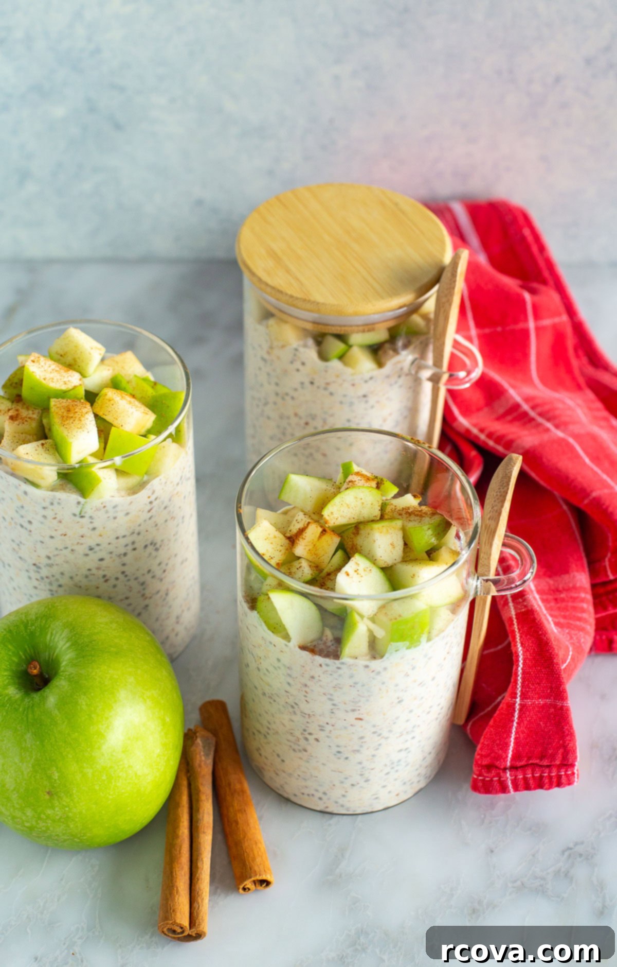 Cozy Apple Cinnamon Overnight Oats 2 Overhead shot of three glass jars filled with homemade Apple Cinnamon Overnight Oats, garnished with a red cloth and fresh cinnamon sticks, ready for a healthy breakfast.
