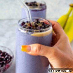Blueberry Tofu Power Smoothie 8 Side shot of a hand holding a purple tofu smoothie topped with blueberries and peanut butter, with two bananas in the background.